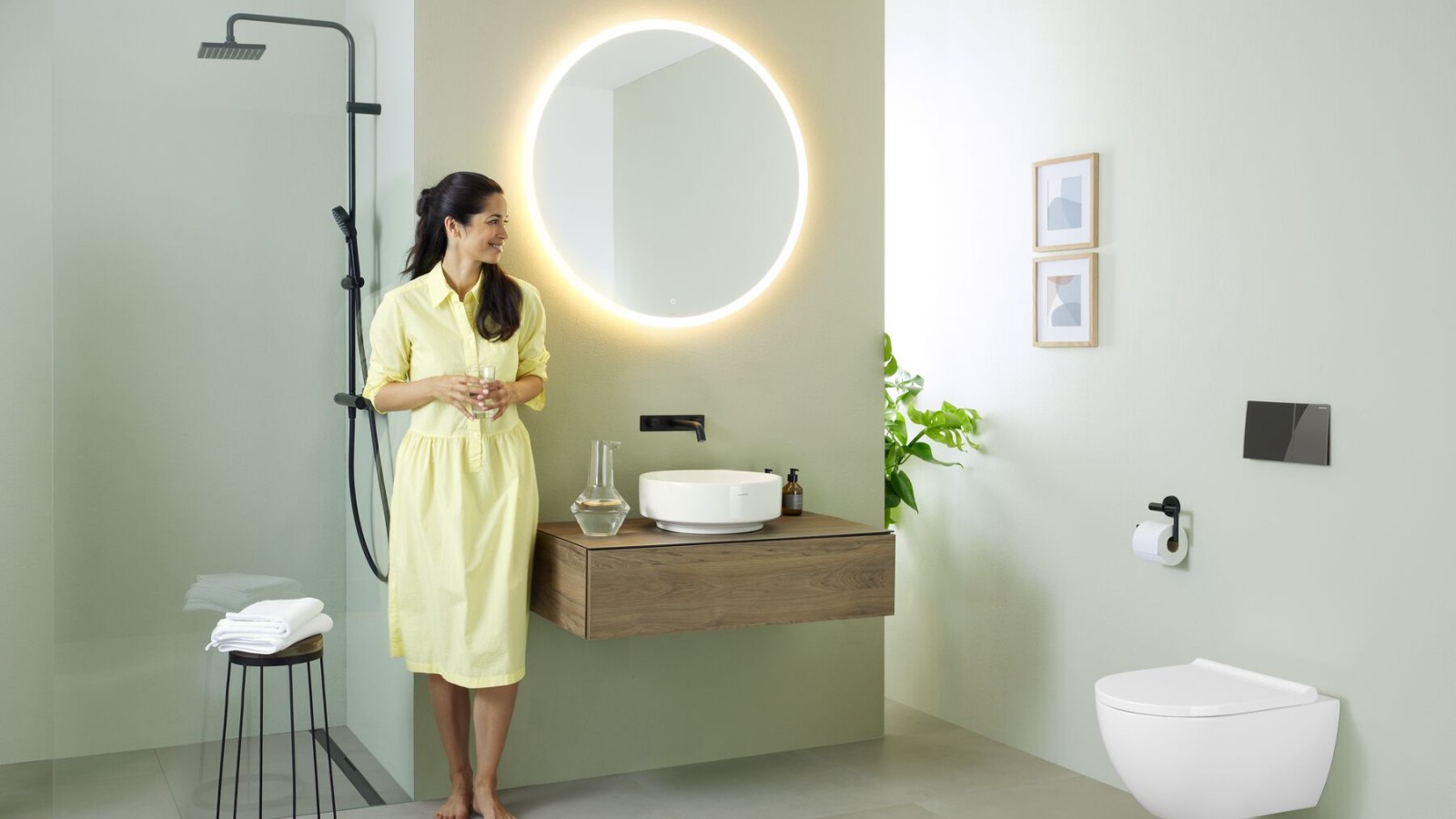 Femme en robe jaune devant une salle de bains vert menthe avec meubles et céramiques de salle de bains Geberit et robinets noirs. Femme en robe jaune devant une salle de bains vert menthe avec meubles et céramiques de salle de bains Geberit et robinets noirs.