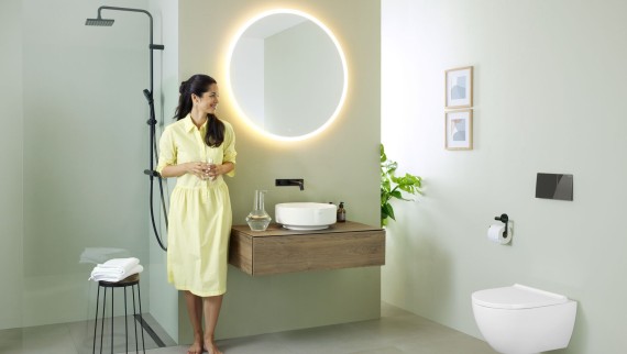 Donna in abito giallo in piedi davanti a un bagno verde menta con mobili e sanitari di Geberit e rubinetti neri Donna in abito giallo in piedi davanti a un bagno verde menta con mobili e sanitari di Geberit e rubinetti neri