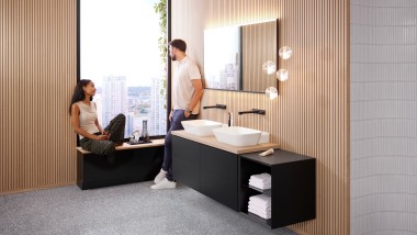 Una donna e un uomo in un grande bagno con lavabo da appoggio Geberit ONE, specchio Option ed elementi in legno