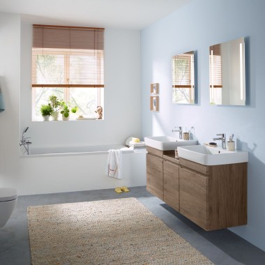 Une salle de bains familiale avec une paroi bleu clair et des meubles de salle de bains en noyer hickory, une armoire de toilette, une plaque de déclenchement et des céramiques de Geberit